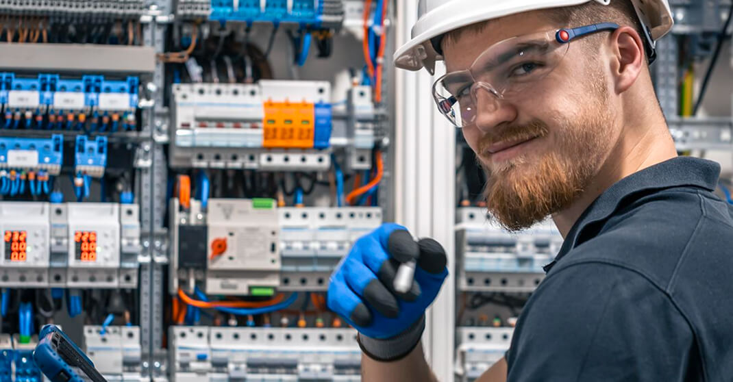 Elektriker mit Schutzhelm und Schutzbrille lächelt vor geöffnetem Schaltschrank