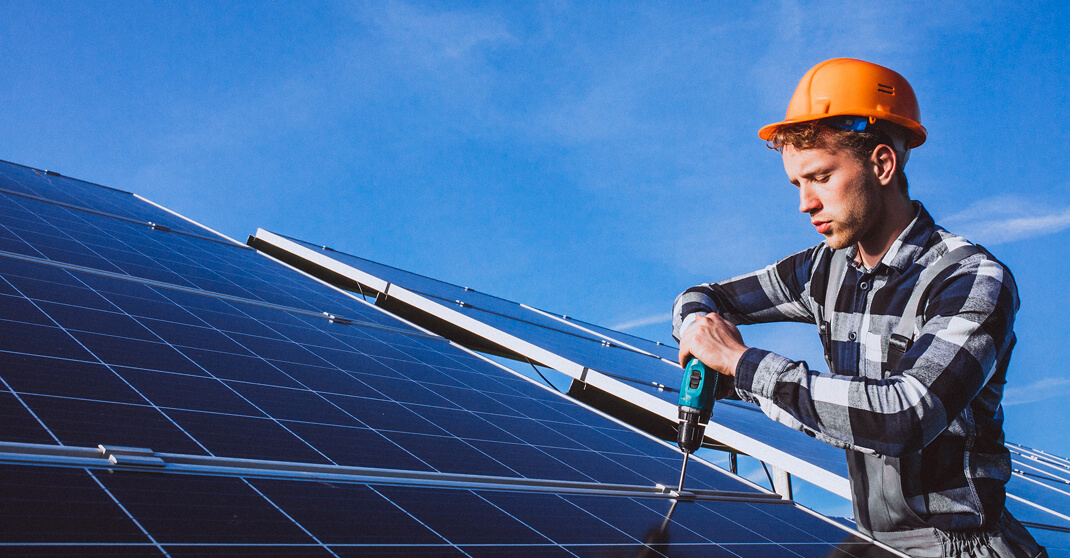 Elektriker mit Schutzhelm sichert Photovoltaikmodul mit Bohrmaschine auf Dach