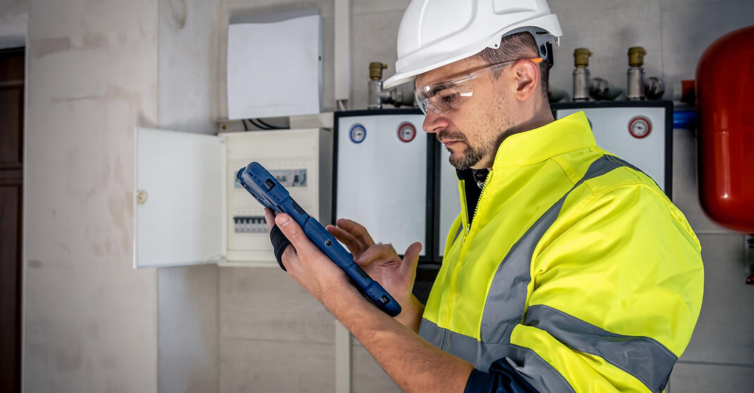 Elektriker mit Schutzhelm prüft Sicherungskasten mit Tablet und Messgerät